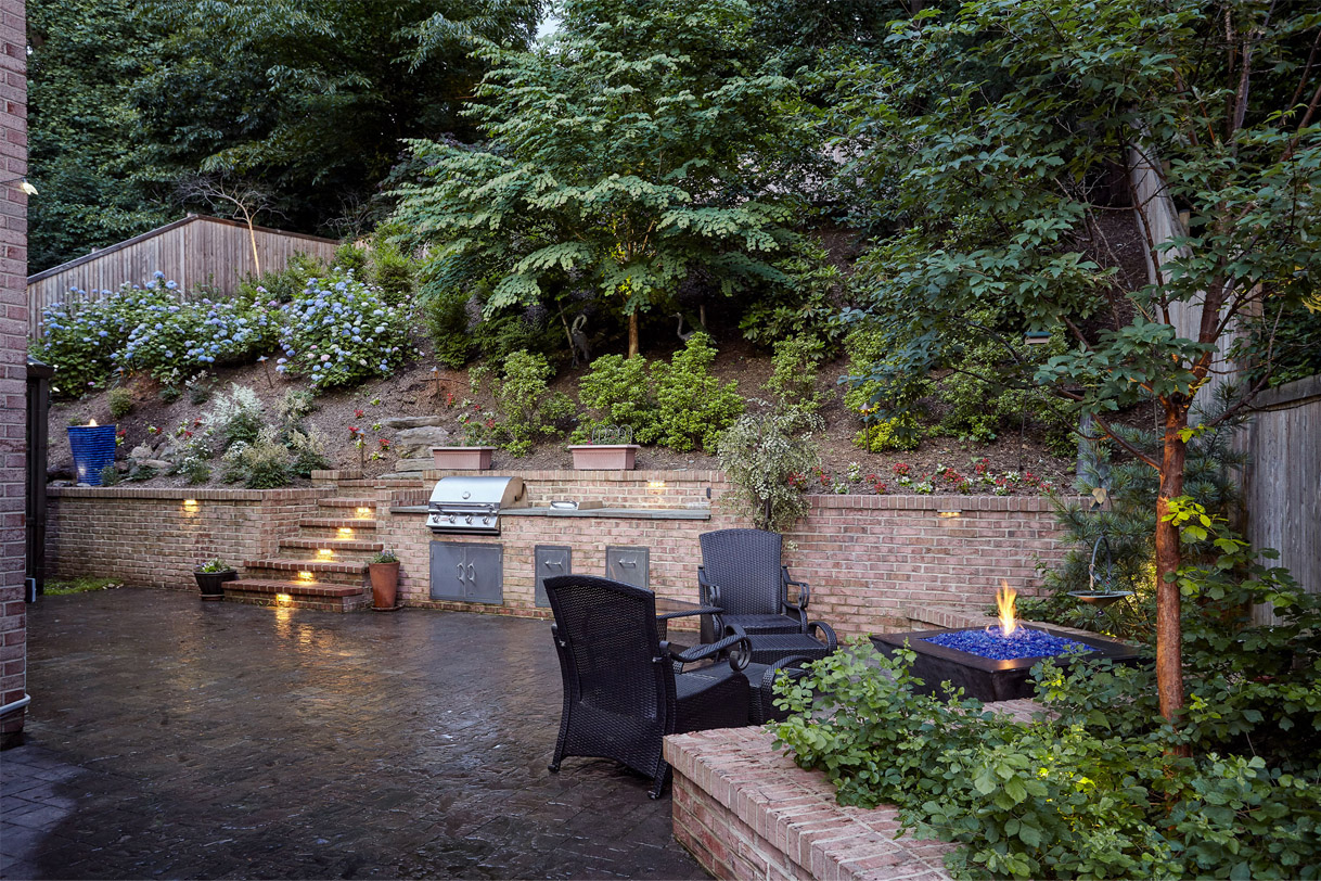 Stone patio with lit fireplace and outdoor lounge furniture at the base of a steep, landscaped hillside