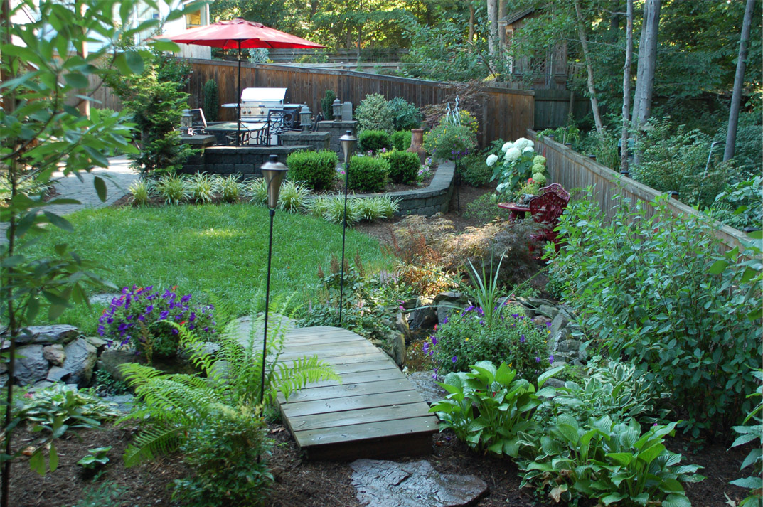 Wooden garden bridge walkway surrounded by lush plantings, connecting lawn to a landscaped patio area with seating and outdoor lighting.