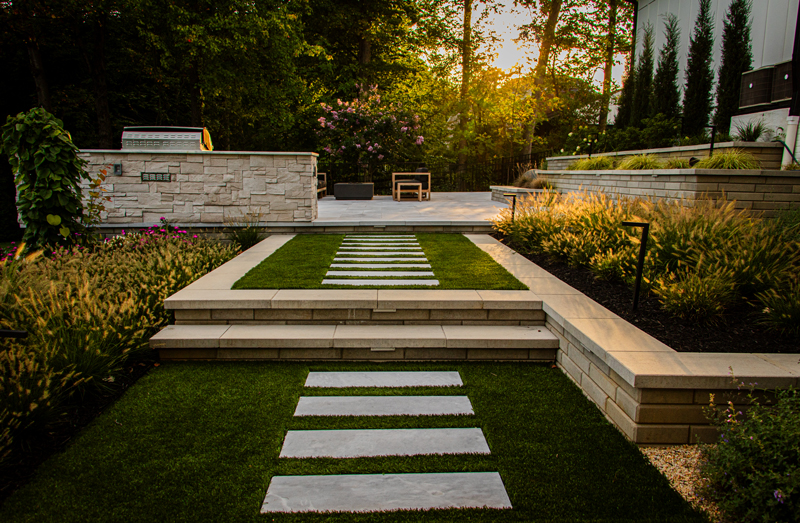 Modern stepping stone walkway set in turf leading to a raised patio with clean-lined stonework and outdoor seating.