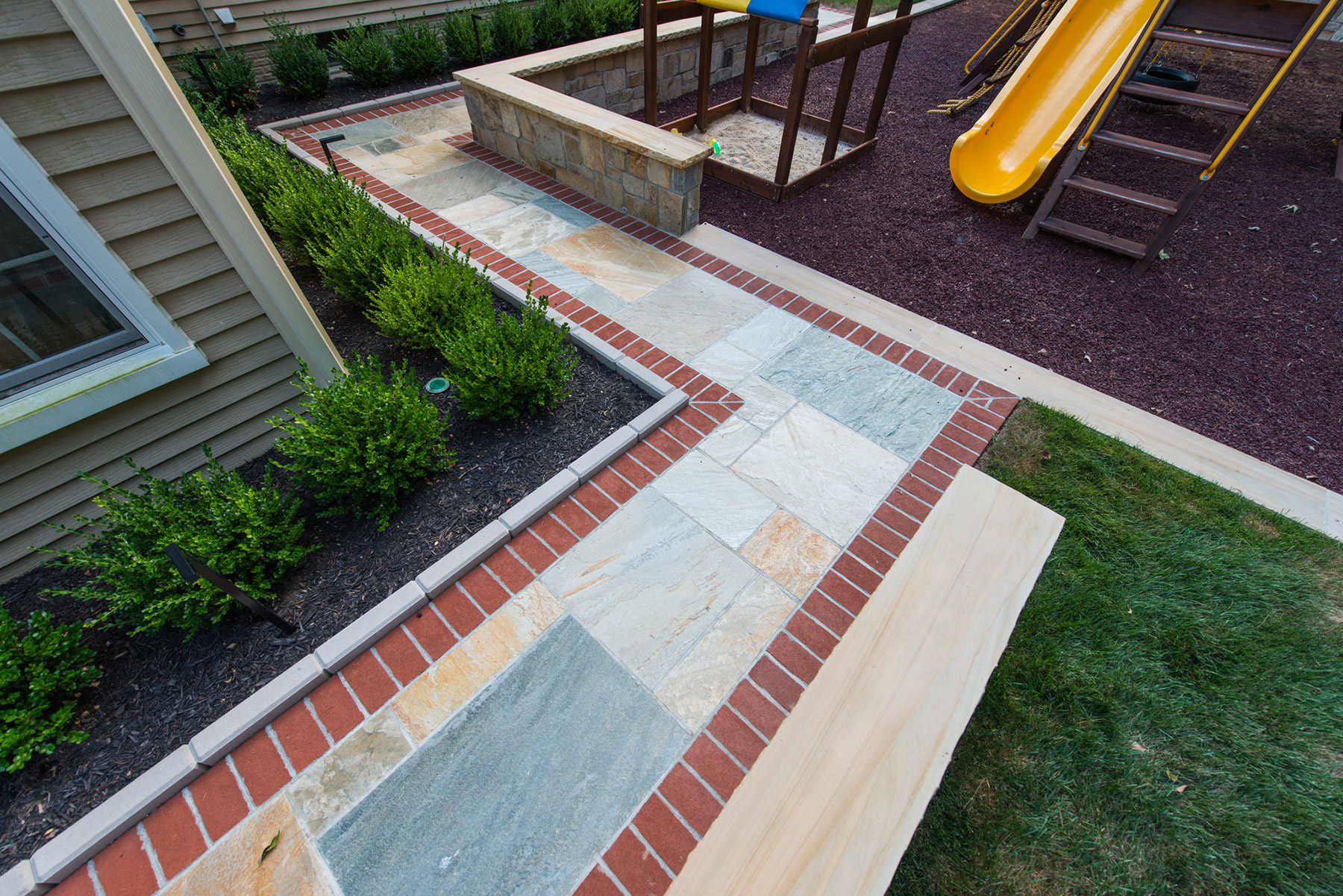 Walkway made of natural stone slabs with red brick borders, leading through a landscaped yard beside a play area and raised planter wall.